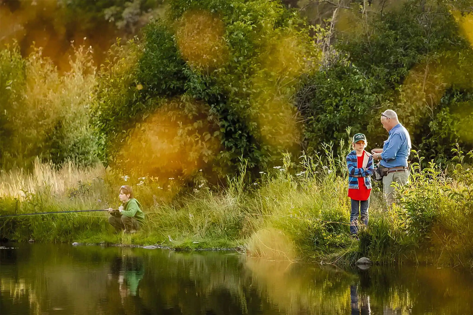 A grand father and two children fly fishing together at the edge of a calm pond at golden hour | Jackson Hole Fly Company