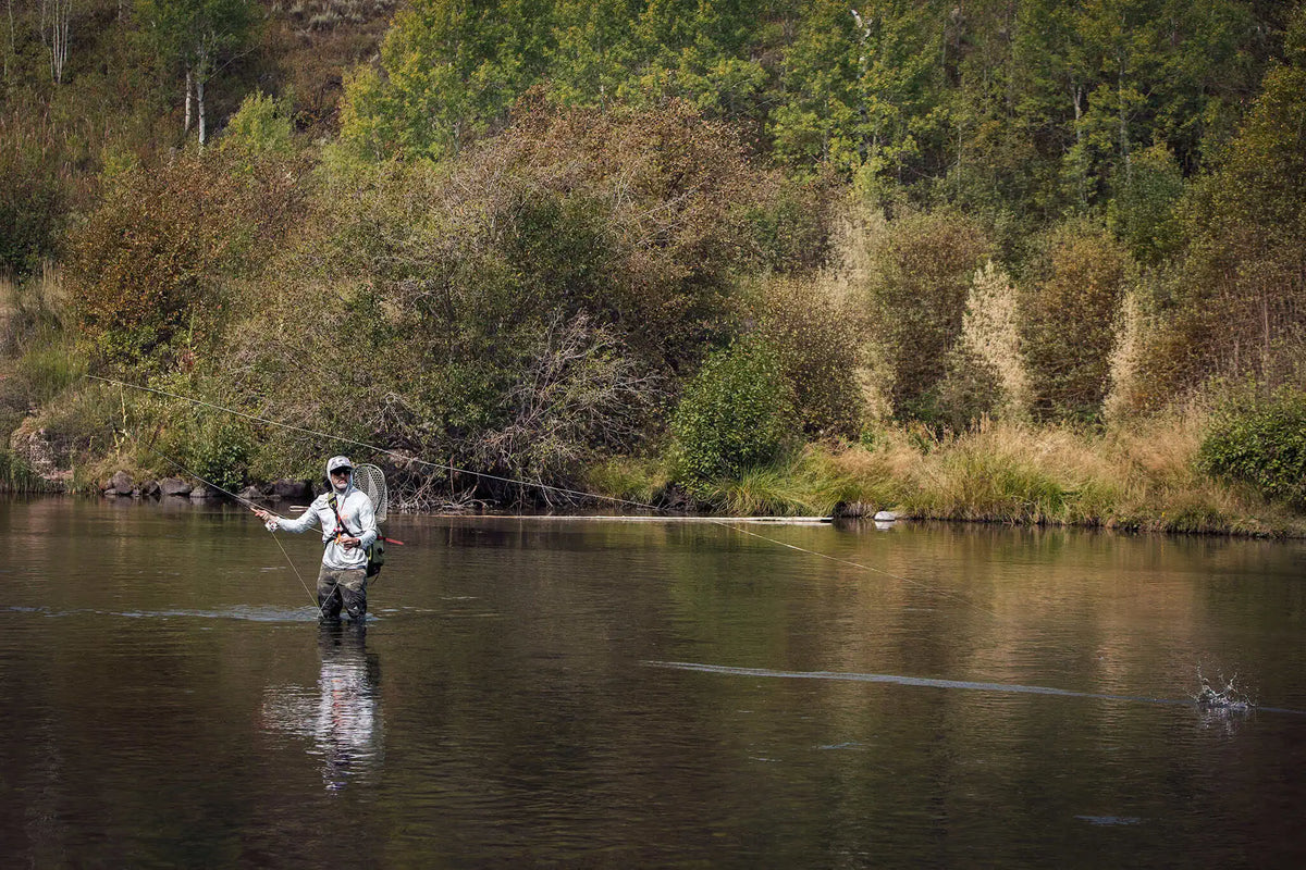 Angler in a JHFLYCO sun hoodie wading a calm trout river, line tight across the current to a splashing fish, landing net on his back | Jackson Hole Fly Company